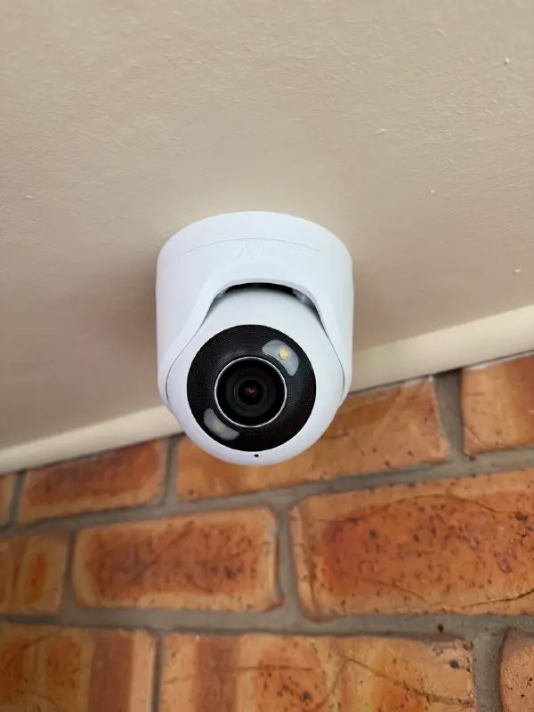 White security camera mounted on a ceiling over a brick wall, angled downward to view the area below.