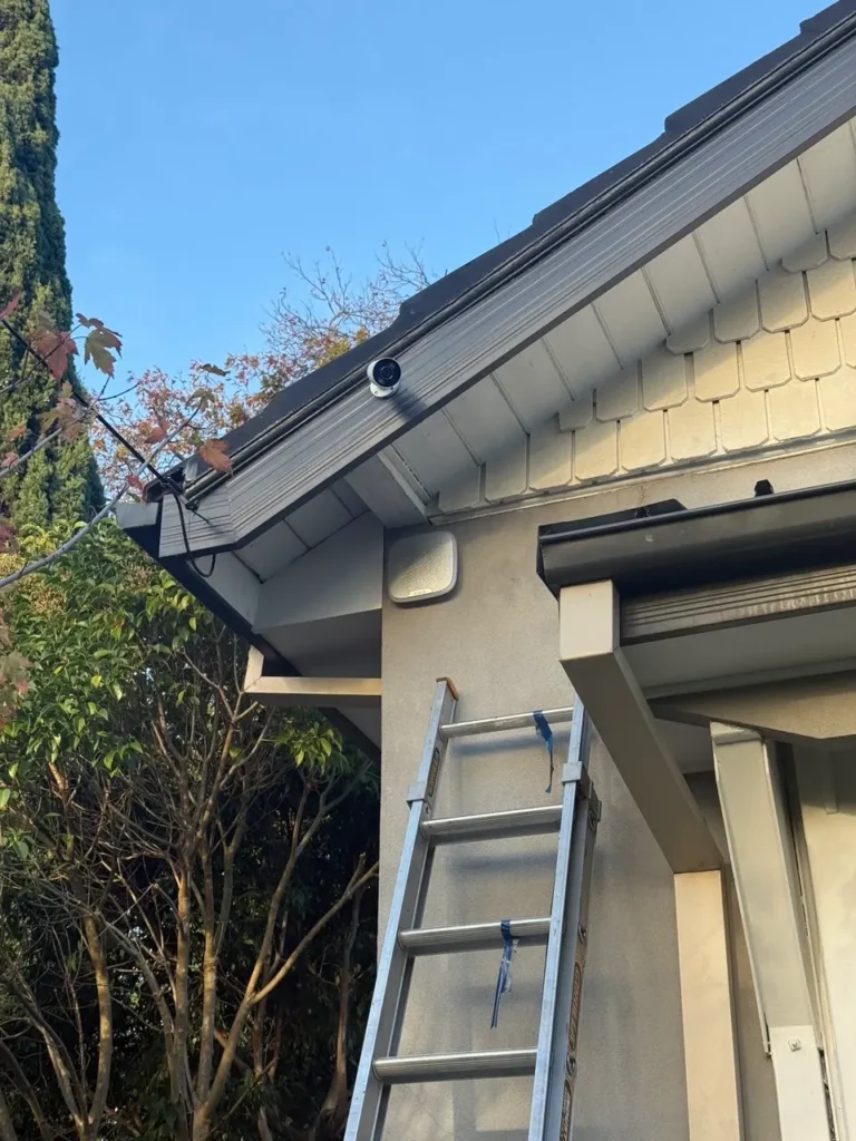 Aluminum ladder leaning against a gray house under a clear blue sky, with a security camera mounted on the roof edge.