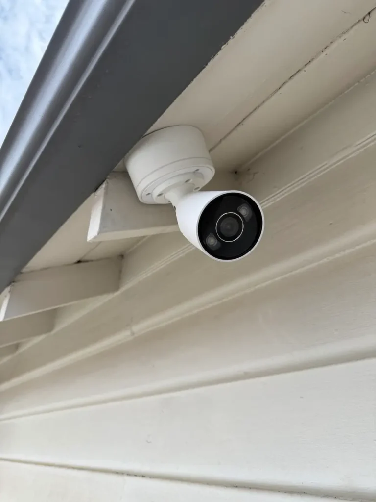Outdoor white security camera mounted under a roof overhang, angled downward for surveillance at the entrance.