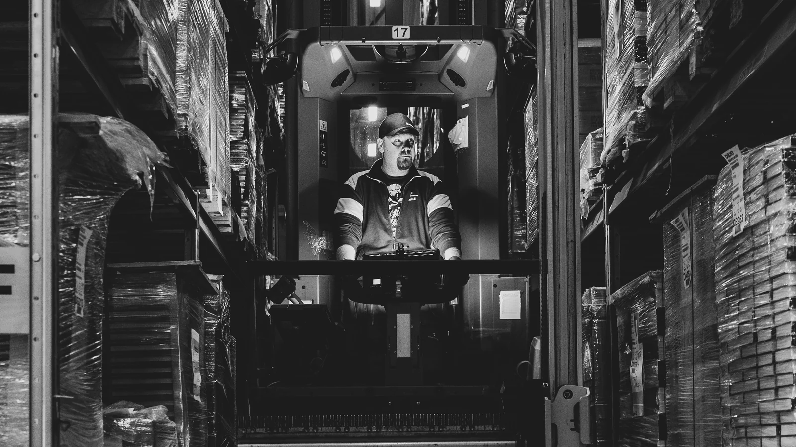 Forklift operator between warehouse racks - SIPKO Security Black-and-white photograph of a forklift operator in a narrow warehouse aisle between stocked pallet racks.