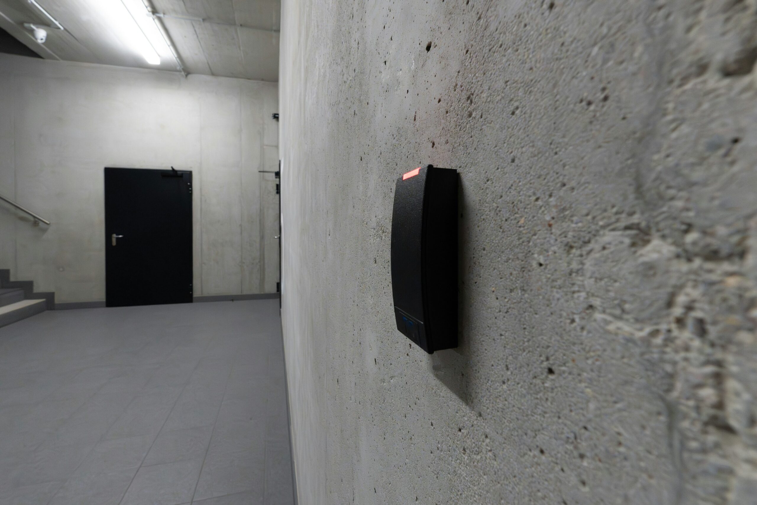Access control card reader for alarm panel comparison - SIPKO Security Modern electronic access card reader mounted on a concrete stairwell wall near a closed door